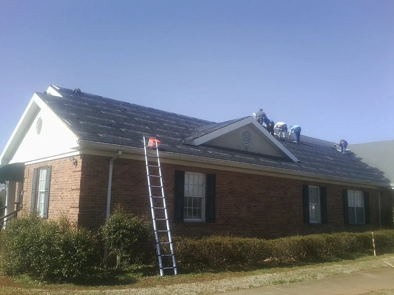 Crew tearing shingles off a brick ranch roof with a ladder set — Emergency Roof Repair work by our crew in North Prairie