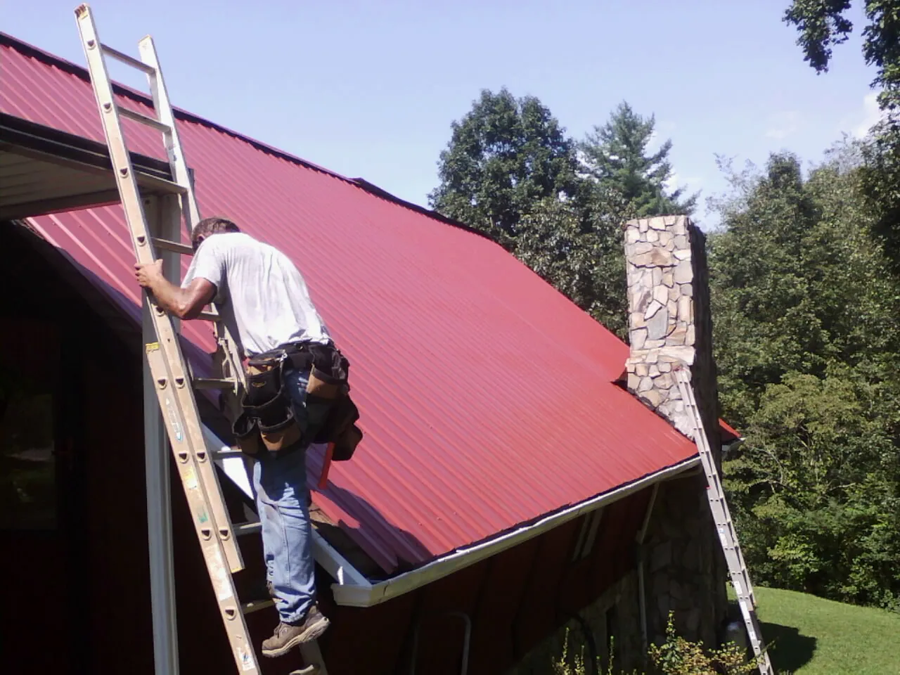Roofer on a ladder beside a red metal roof and stone chimney — Soffit & Fascia Repair by Roofing Brothers in North Prairie